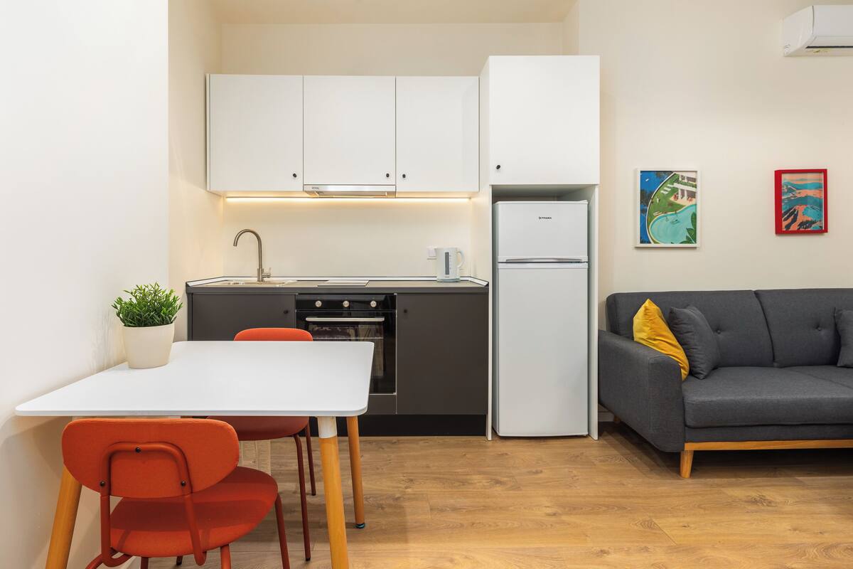 Una cucina compatta con mobili bianchi, un frigorifero e una zona pranzo con tavolo bianco e sedie arancioni.