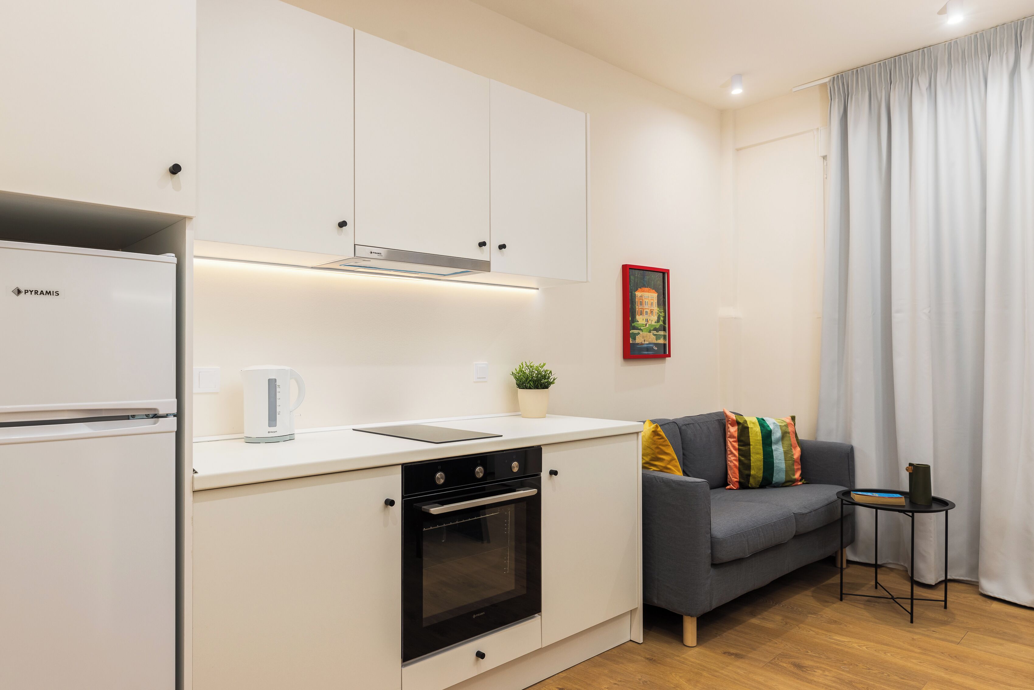 A compact kitchen with white cabinets, a built-in oven, and a coffee maker. There's a gray sofa with colorful pillows and a small side table with a plant.