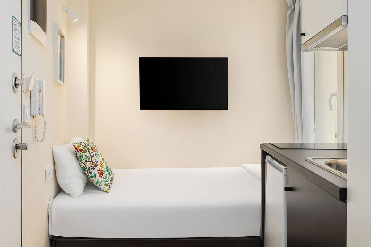 Una stanza compatta con un letto, un angolo cottura, una TV a parete e un pavimento in legno.