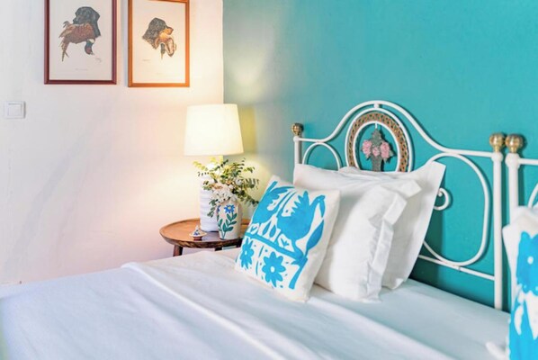 A bed with white bedding and a teal headboard, a bedside table with a lamp and a vase of flowers, and two framed pictures on the wall.