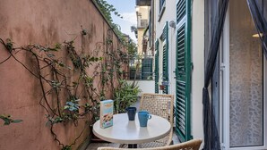 Une petite table ronde, sur laquelle sont posées deux tasses bleues et une boîte, est entourée de chaises en osier. L’ensemble se trouve dans un petit espace extérieur, bordé d’un mur rouge et agrémenté de volets verts.
