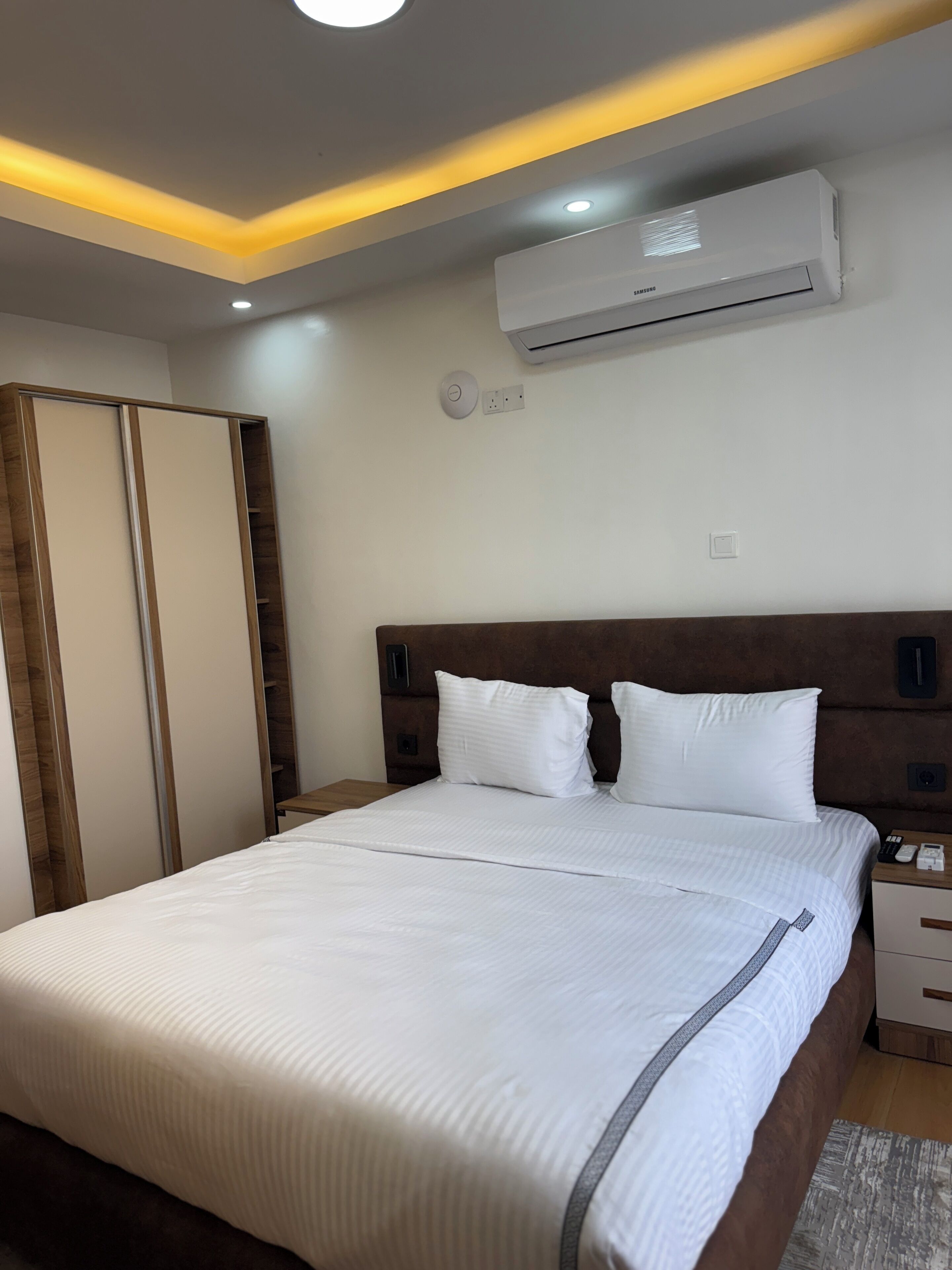 A modern hotel room with a large bed, wooden headboard, bedside tables, an air conditioning unit, and a ceiling light fixture.