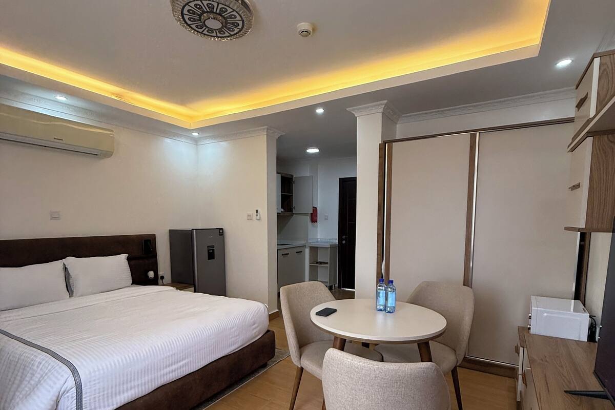 Une chambre d’hôtel moderne dotée d’un grand lit, d’une table de salle à manger ronde avec des chaises, d’une kitchenette et d’un plafond équipé de luminaires encastrés.
