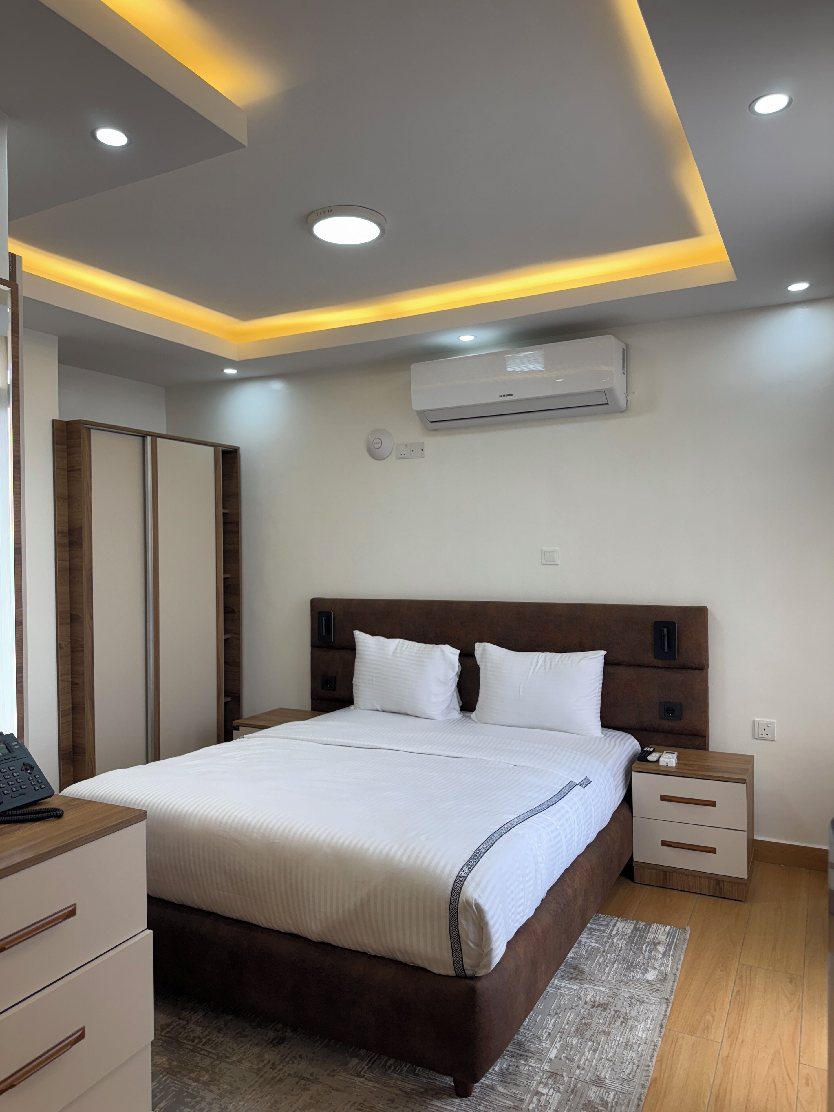 A modern bedroom with a large bed, bedside tables, a wardrobe, and an air conditioning unit.