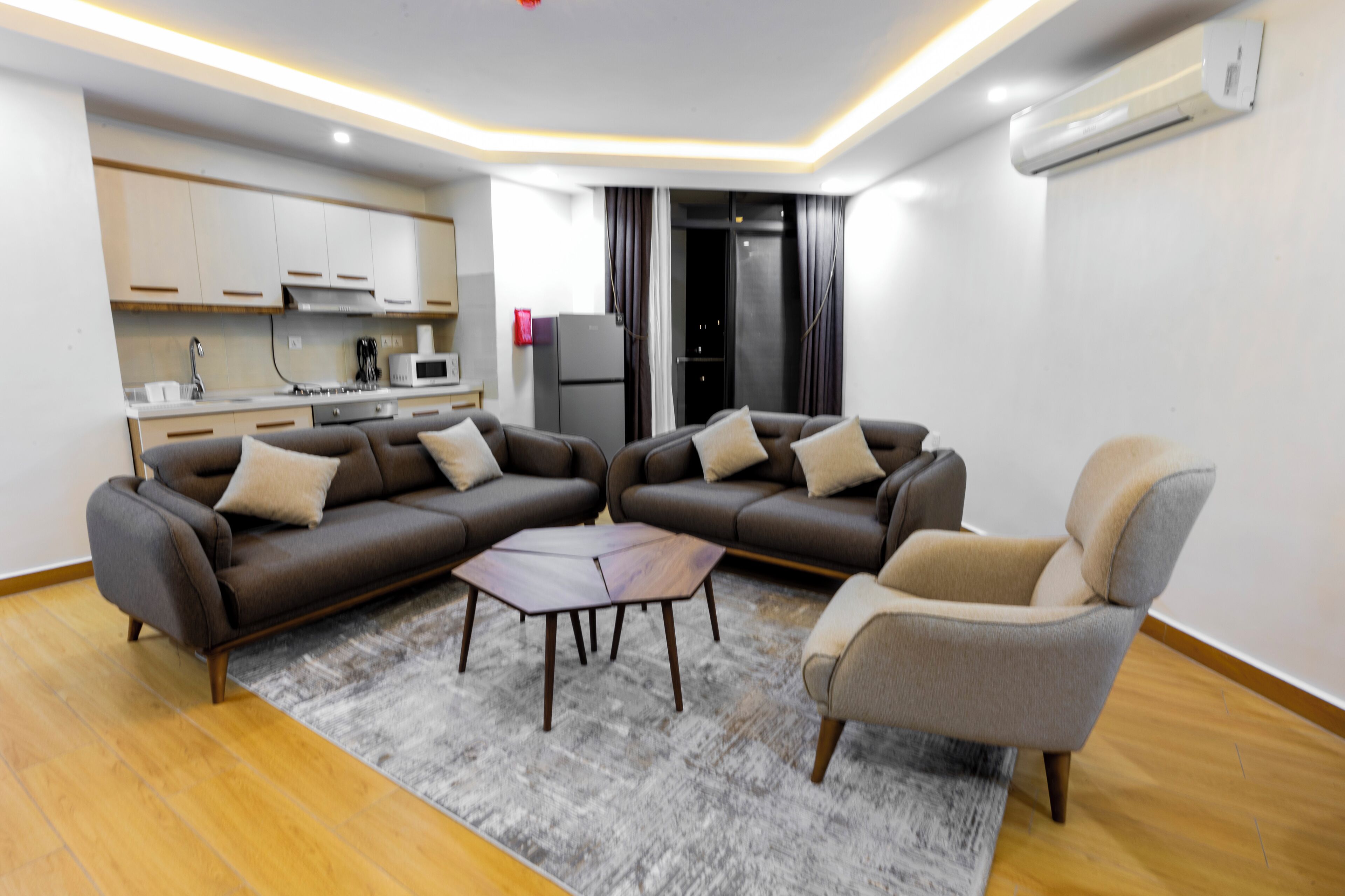 A modern living room with a sofa set, a coffee table, and a chair. There is a kitchen area with cabinets and appliances in the background.