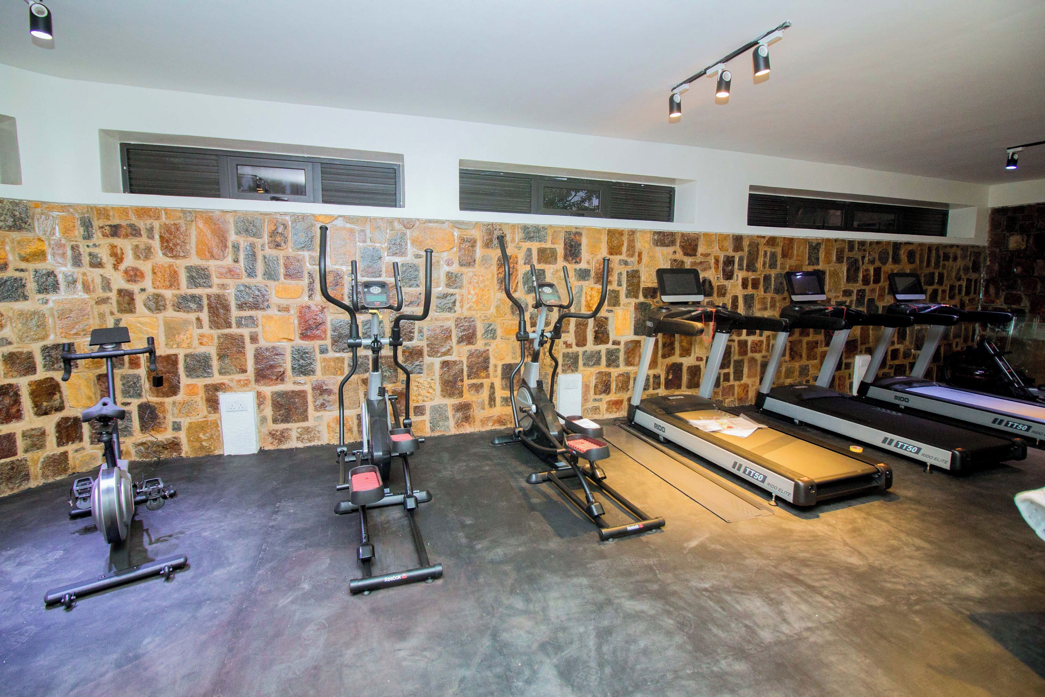 Een fitnessruimte met verschillende cardio-apparaten, een stenen muur en een verlaagd plafond met verlichting.