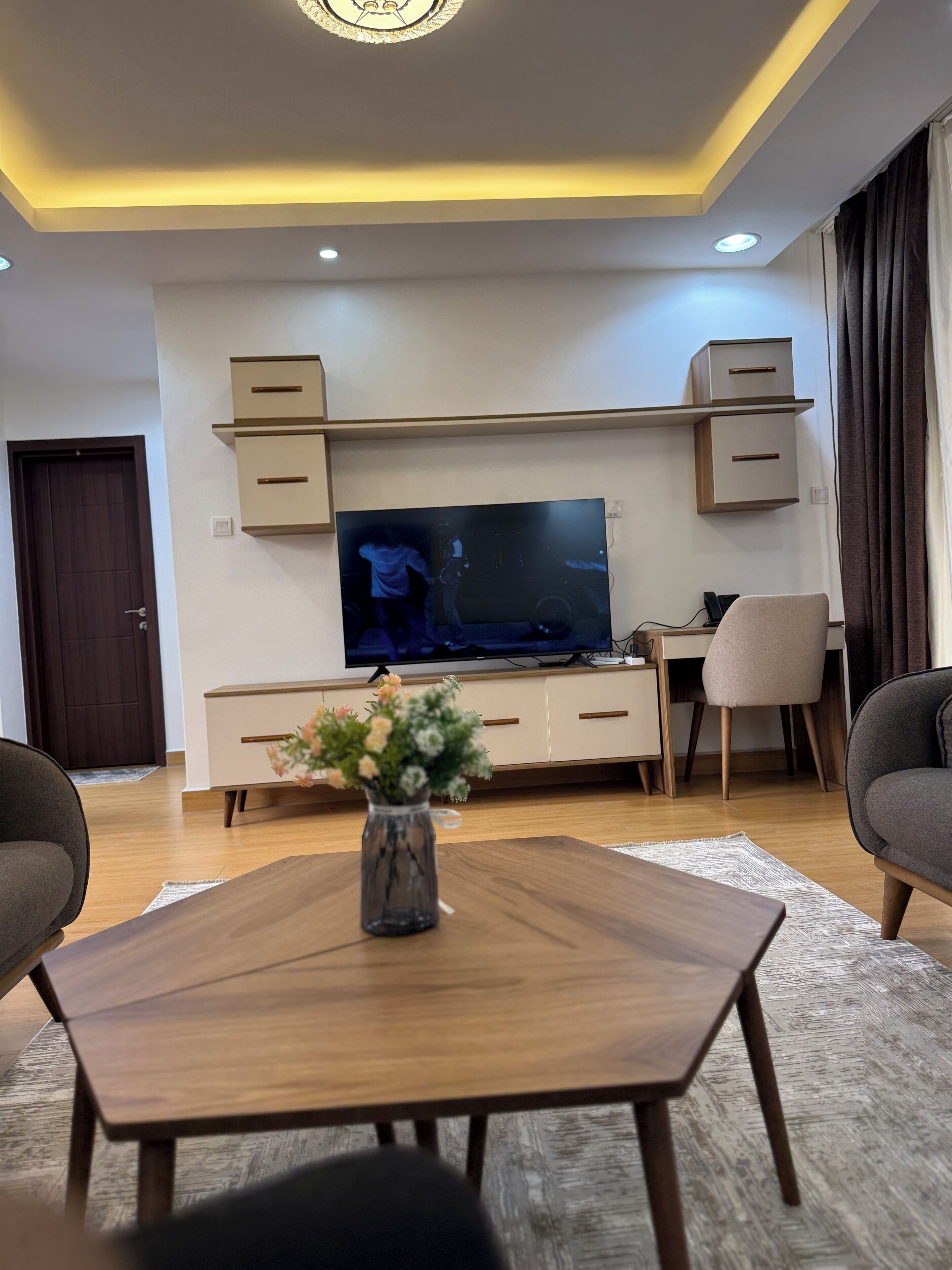 A modern living room with a flat-screen TV, wooden coffee table, and a vase of flowers.