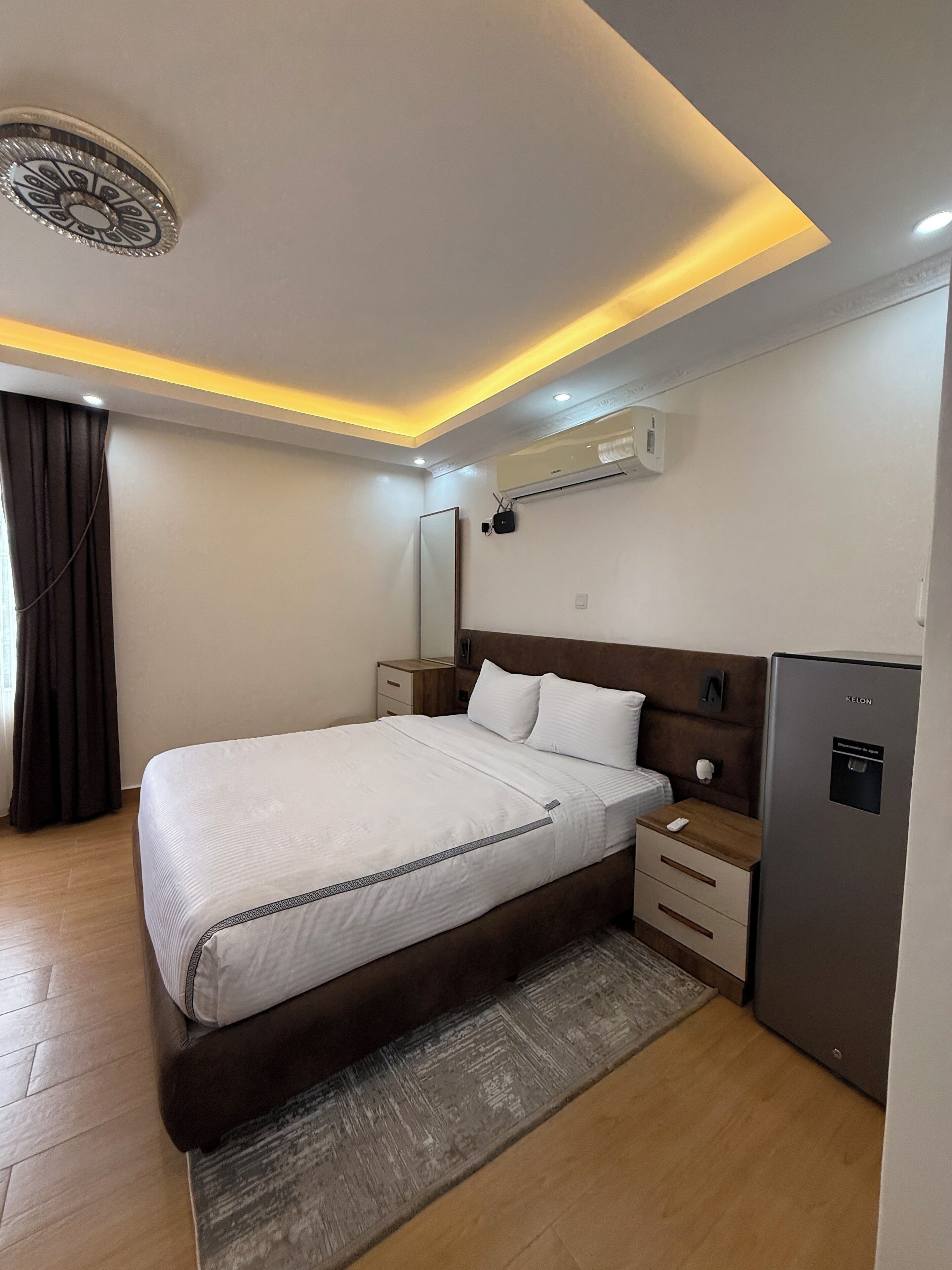 Habitación de hotel con una cama grande, mesitas de noche, un minibar, un equipo de aire acondicionado e iluminación en el techo.