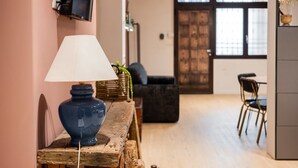 Une table en bois avec une lampe et un vase, un téléviseur fixé au mur, et un coin repas avec des chaises et une table à l’arrière-plan.