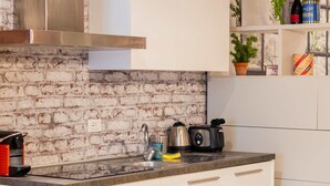 Une cuisine avec des armoires blanches, un mur en briques, une hotte de cuisinière en acier inoxydable, un évier, un plan de travail avec une bouilloire et un grille-pain, et un coin repas avec une chaise et une table.
