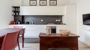 A modern kitchen with white cabinets, a dark countertop, and a wooden dining table with chairs. There are framed pictures on the wall above the countertop. - Casa Melzo Chic Apartment in Porta Venezia (Milan)