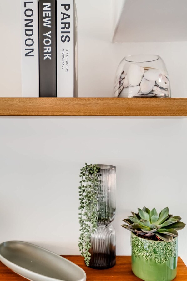 Una estantería con libros titulados 'LONDON', 'NEW YORK' y 'PARIS', un florero de vidrio con guijarros blancos, una planta verde y una bandeja metálica.