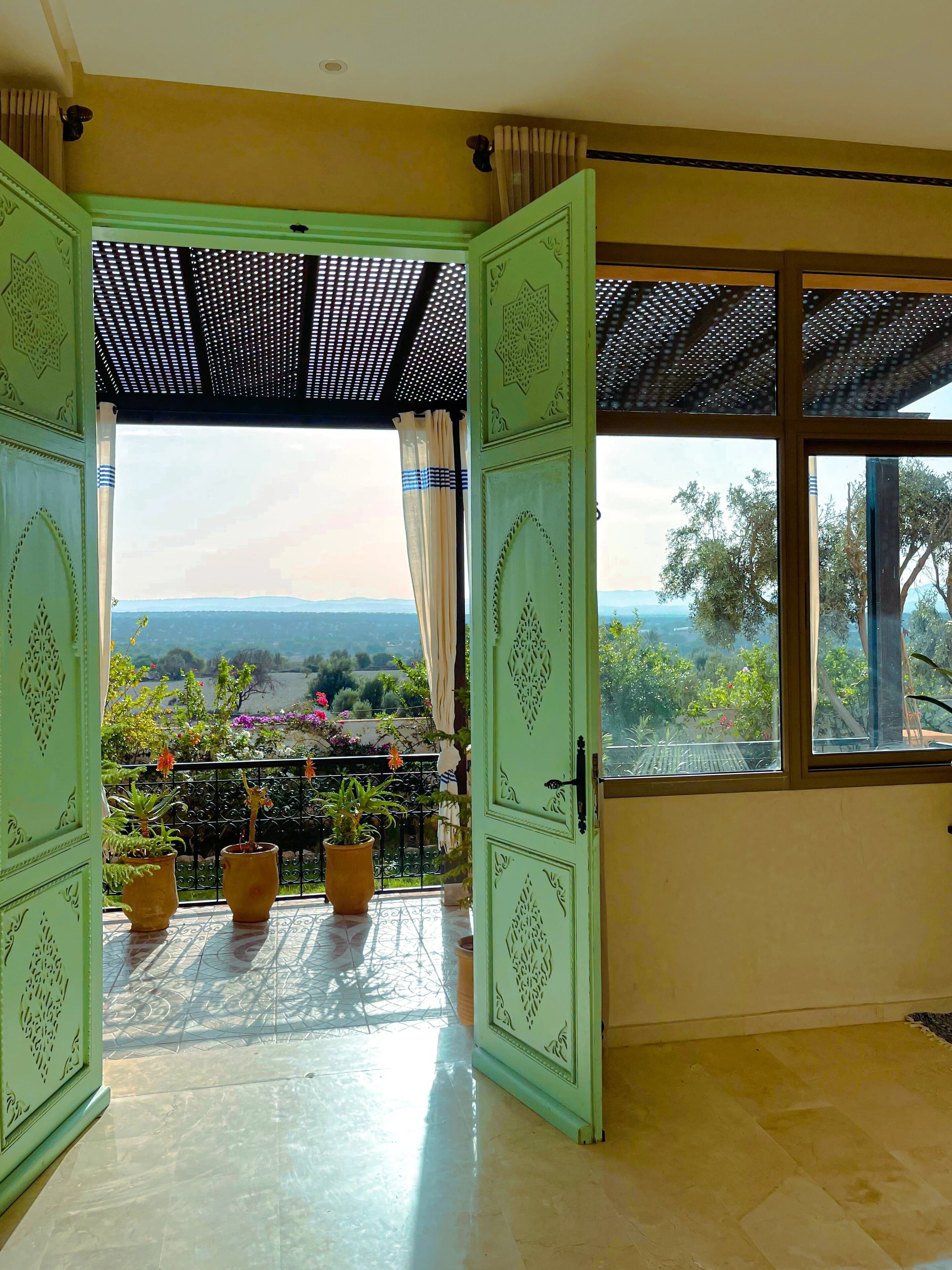 Une porte ouverte donnant sur un balcon agrémenté de plantes en pot, d’où l’on peut admirer le paysage, et sur lequel se trouve une pergola.