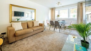 Ein Wohnzimmer mit einem beigen Sofa, einem Couchtisch, einem Essbereich mit Stühlen, einem großen Spiegel und einer Topfpflanze.