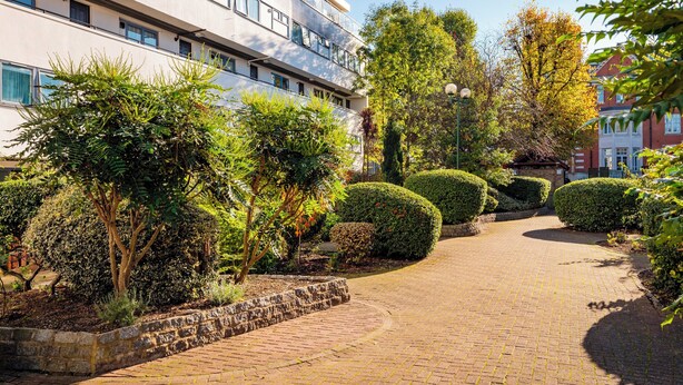 Ein gepflasterter Weg führt zu einem Gebäude mit vielen Fenstern und einem gepflegten Garten mit verschiedenen Sträuchern und Bäumen.