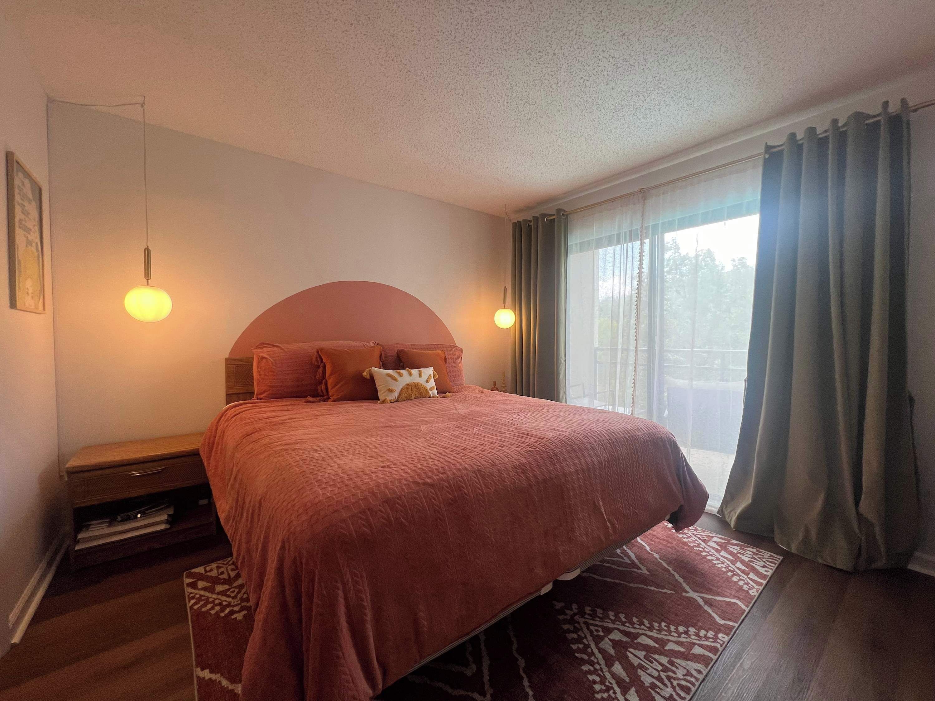 Ein Schlafzimmer mit einem großen Bett, zwei Nachttischlampen, einem Holznachttisch und Blick durch ein mit Vorhängen drapiertes Fenster.