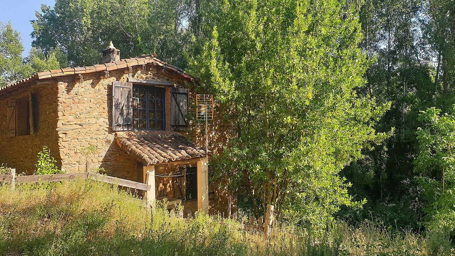 A rustic stone building with a tiled roof, surrounded by dense greenery.