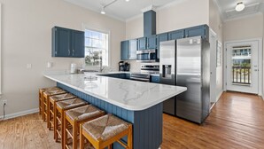 A modern kitchen with blue cabinets, a large island with bar stools, stainless steel appliances, and a view of the outdoors. - Sunflower Mama, a gulf front home. (St. George Island)