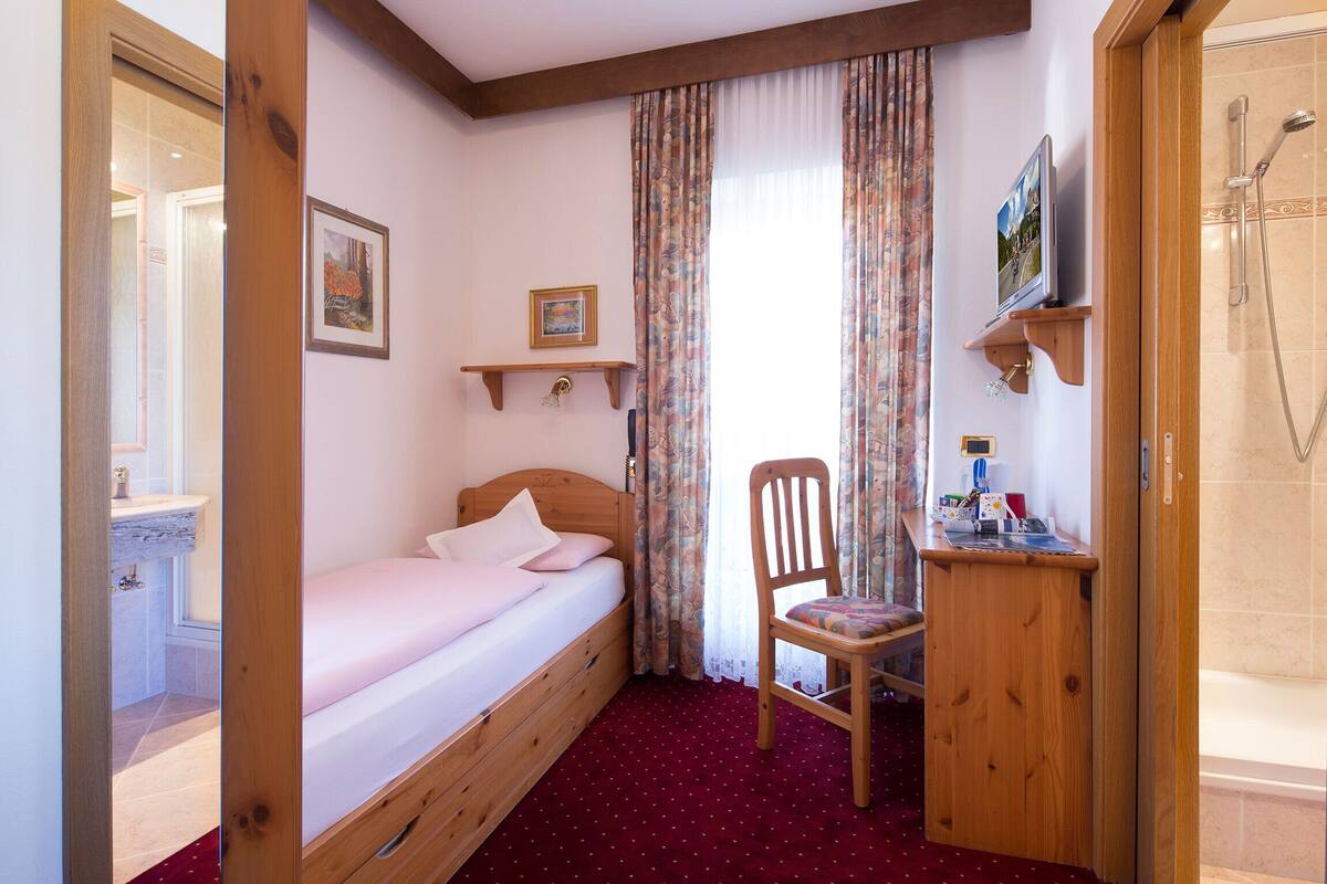 A bedroom with a single bed, a wooden chair, a small desk with a mirror, a shower area, and a window with curtains.