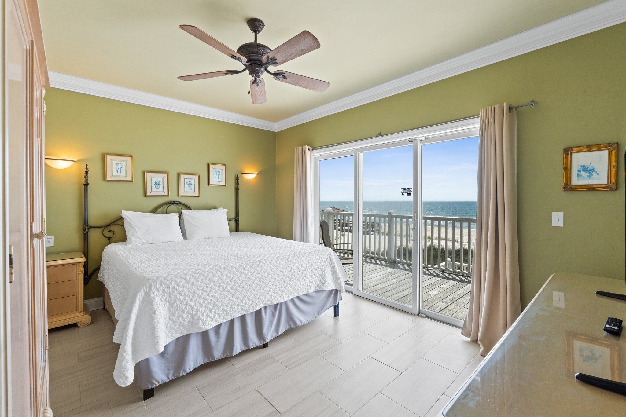 A bedroom with a bed, a ceiling fan, a nightstand, a desk, and a view of the ocean through a sliding glass door.