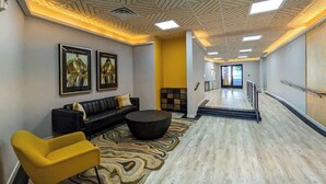 Eine moderne Lobby mit einem schwarzen Ledersofa, einem gelben Sessel und einem runden schwarzen Couchtisch. An den Wänden befinden sich gerahmte Kunstwerke und eine Treppe, die zu einer anderen Ebene führt.