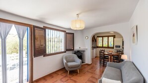A compact living space with a sofa, armchair, and dining table. A fireplace is visible in the background, and a kitchen area is seen through an archway.