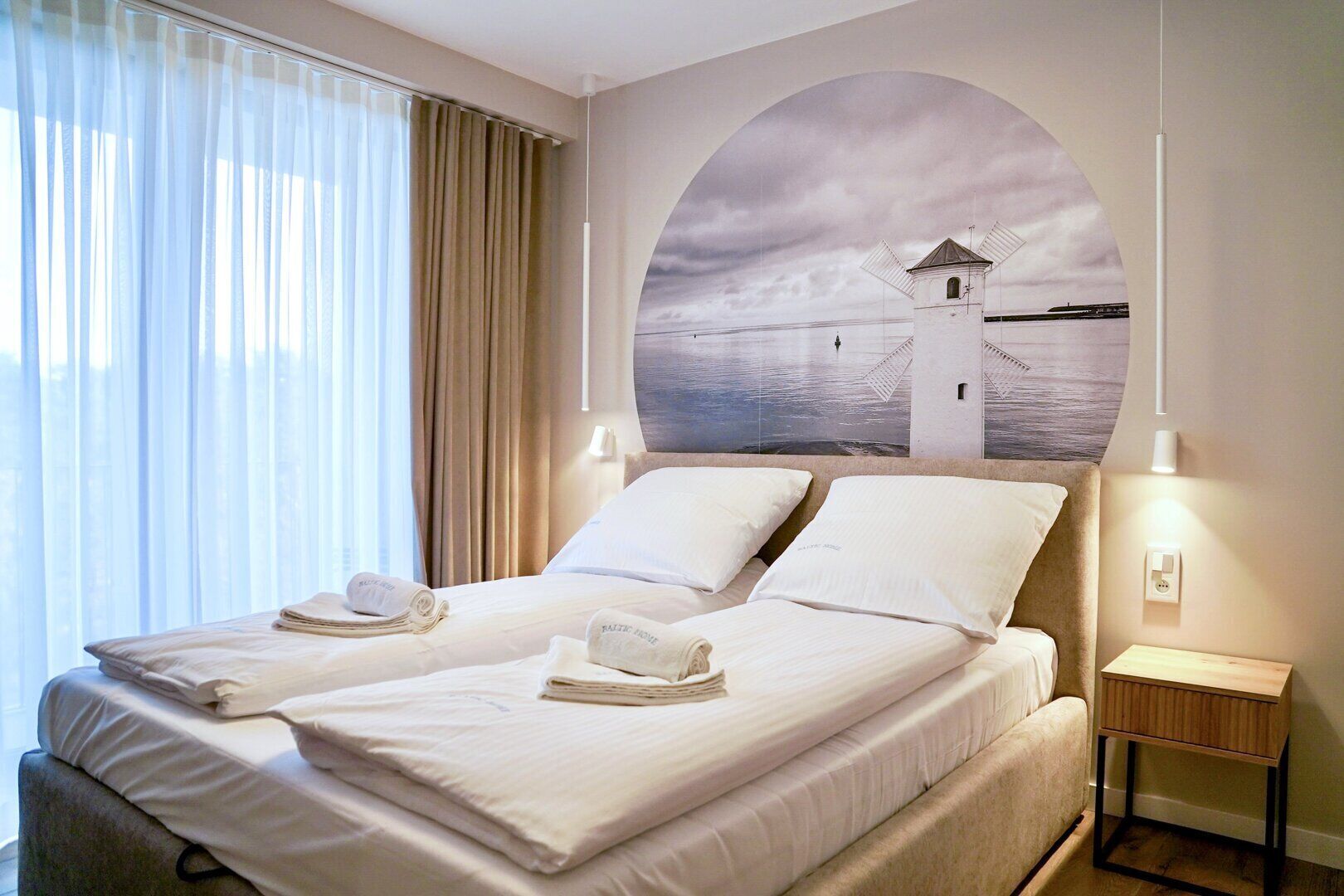 A hotel room with a bed, two pillows, a folded towel, a nightstand, and a wall-mounted lamp.