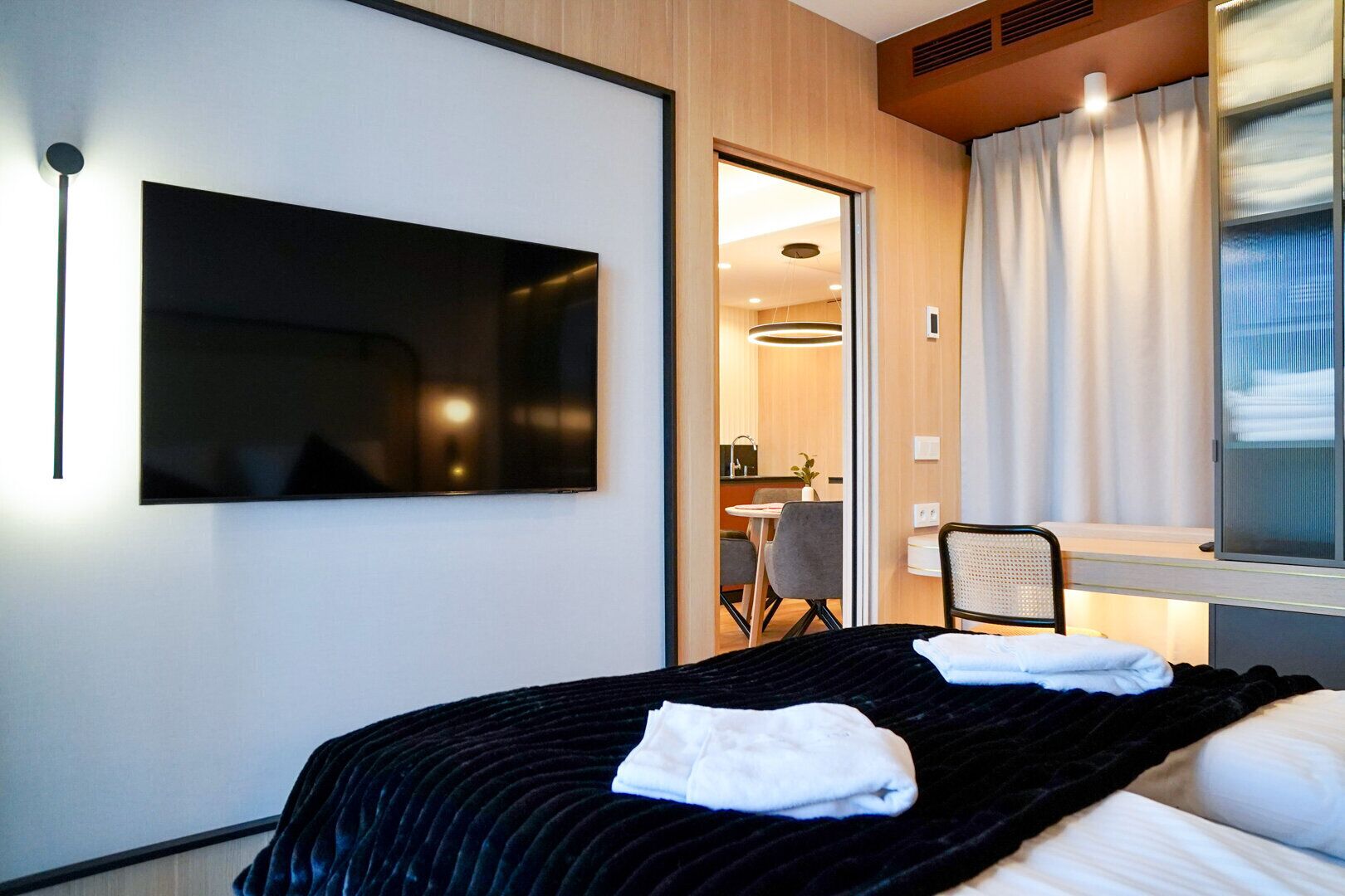A hotel room with a large bed, a flat-screen TV mounted on the wall, a small desk with a chair, and a window with curtains.
