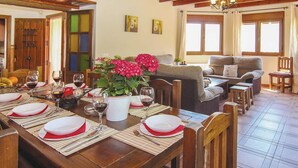 Uma sala de jantar com uma mesa de madeira posta para refeição, uma planta em vaso com flores vermelhas, e uma sala de estar com um sofá e uma janela.