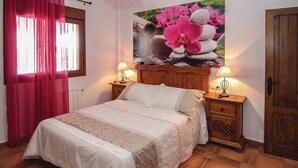 Quarto com cama, duas mesas de cabeceira, cabeceira de madeira, janela com cortinas e um quadro de flores na parede.