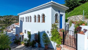 A white building with multiple windows and a blue door, surrounded by potted plants and a stone wall. - Frigiliana 029 (Frigiliana)