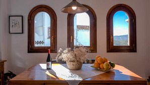 A wooden table with a bottle of wine, a basket of dried flowers, and a plate of fruit. Three arched windows with views of a building and landscape are visible. - Frigiliana 029 (Frigiliana)