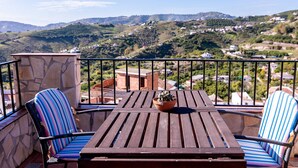 A balcony with a wooden table and two striped chairs, a potted plant on the table, and a stone wall. - Frigiliana 029 (Frigiliana)