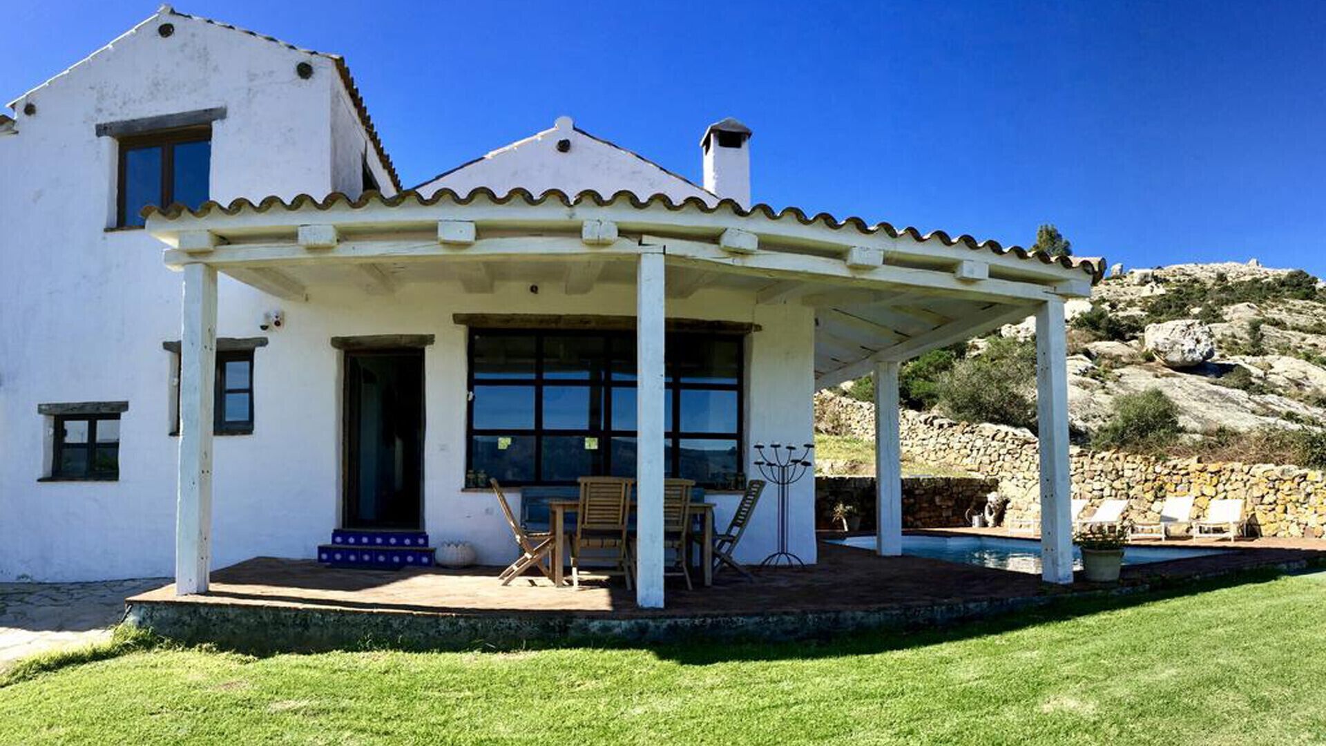 A white building with a covered patio, a pool, and a mountainous backdrop.
