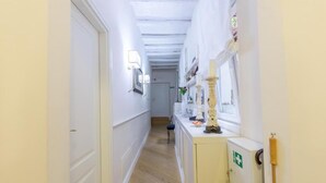 Un couloir étroit aux murs blancs et au plancher en bois. On y trouve une armoire blanche sur laquelle est apposé un panneau vert indiquant les issues de secours, ainsi qu’une fenêtre ornée d’un rideau blanc.