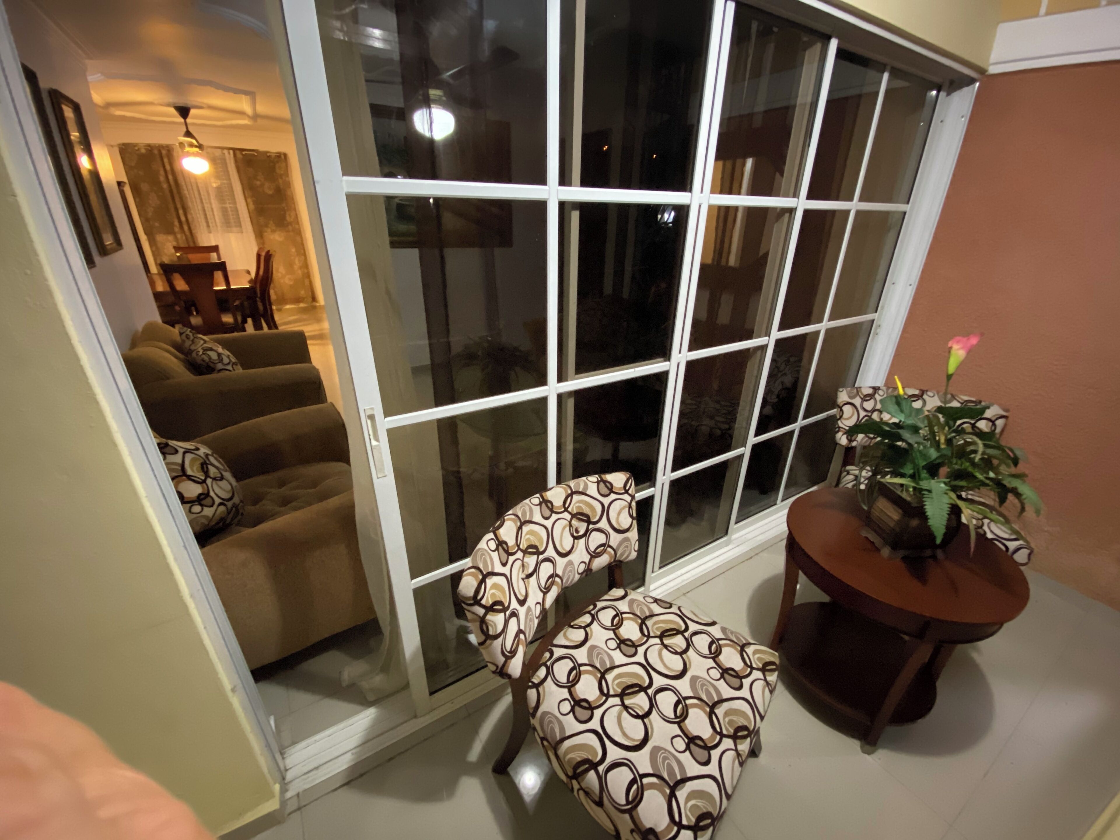 Ein Wohnzimmer mit einer Schiebetür zu einem Balkon. Es gibt eine Sofagarnitur, einen gemusterten Hocker und einen kleinen Tisch mit einer Pflanze.