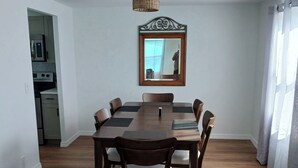 Un comedor con mesa y sillas de madera, un espejo en la pared y una cocina visible a través de una puerta abierta.