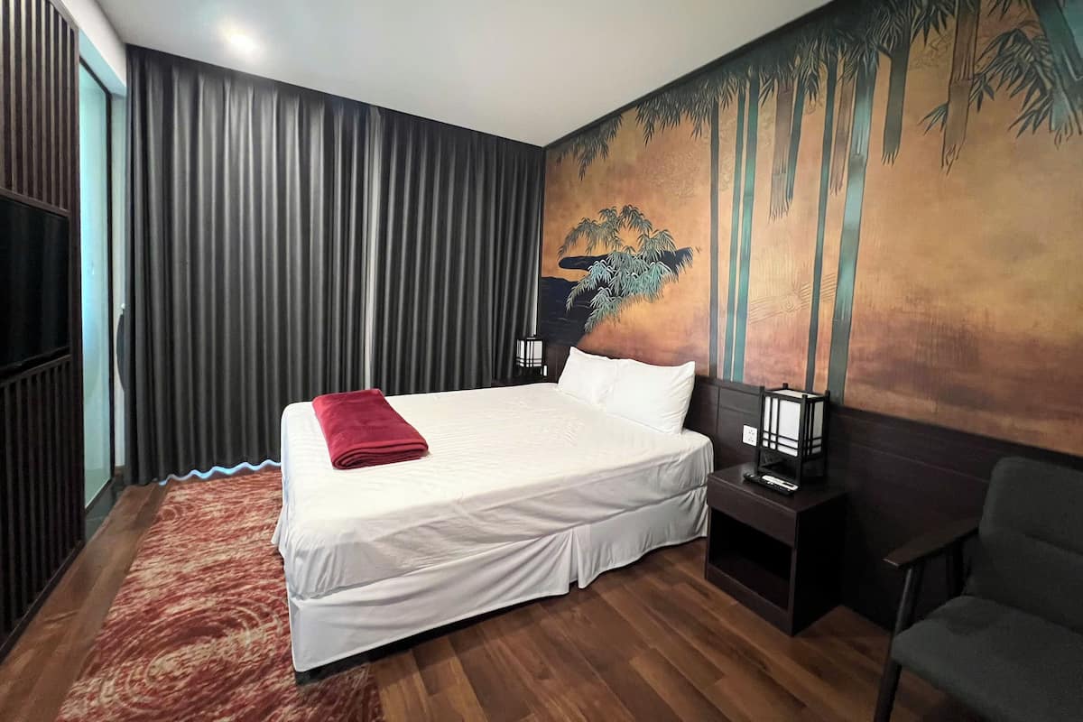 A hotel room with a large bed, a red throw blanket, a nightstand with a lamp, a chair, and a mural of bamboo on the wall.