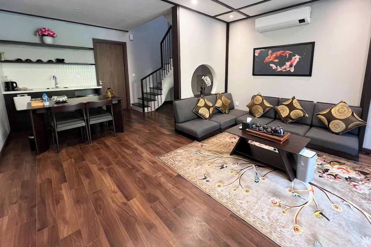A modern living room with a grey sofa, a coffee table, and a rug. There is a kitchen area with a sink and cabinets, and a staircase leading to another level.