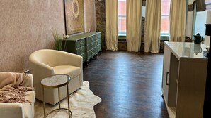 Ein Wohnzimmer mit einem beigen Sessel, einem kleinen runden Tisch, einer grünen Kommode und einem großen Gemälde an der Wand.