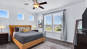 Une chambre à coucher comprenant un lit, un ventilateur de plafond, une fenêtre avec des rideaux, une table de chevet et une commode.
