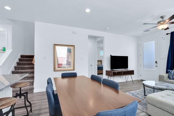 Una sala de estar moderna con una mesa de comedor de madera, sillas tapizadas en azul, un televisor de pantalla plana y un ventilador de techo.