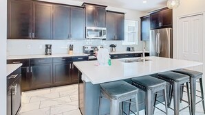 Una cocina moderna con armarios oscuros, electrodomésticos de acero inoxidable, una isla blanca con taburetes e iluminación colgante.