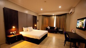A hotel room with a large bed, bedside tables, a desk, a chair, a TV, and a window with curtains. - Genesis Hotels (Port Harcourt)