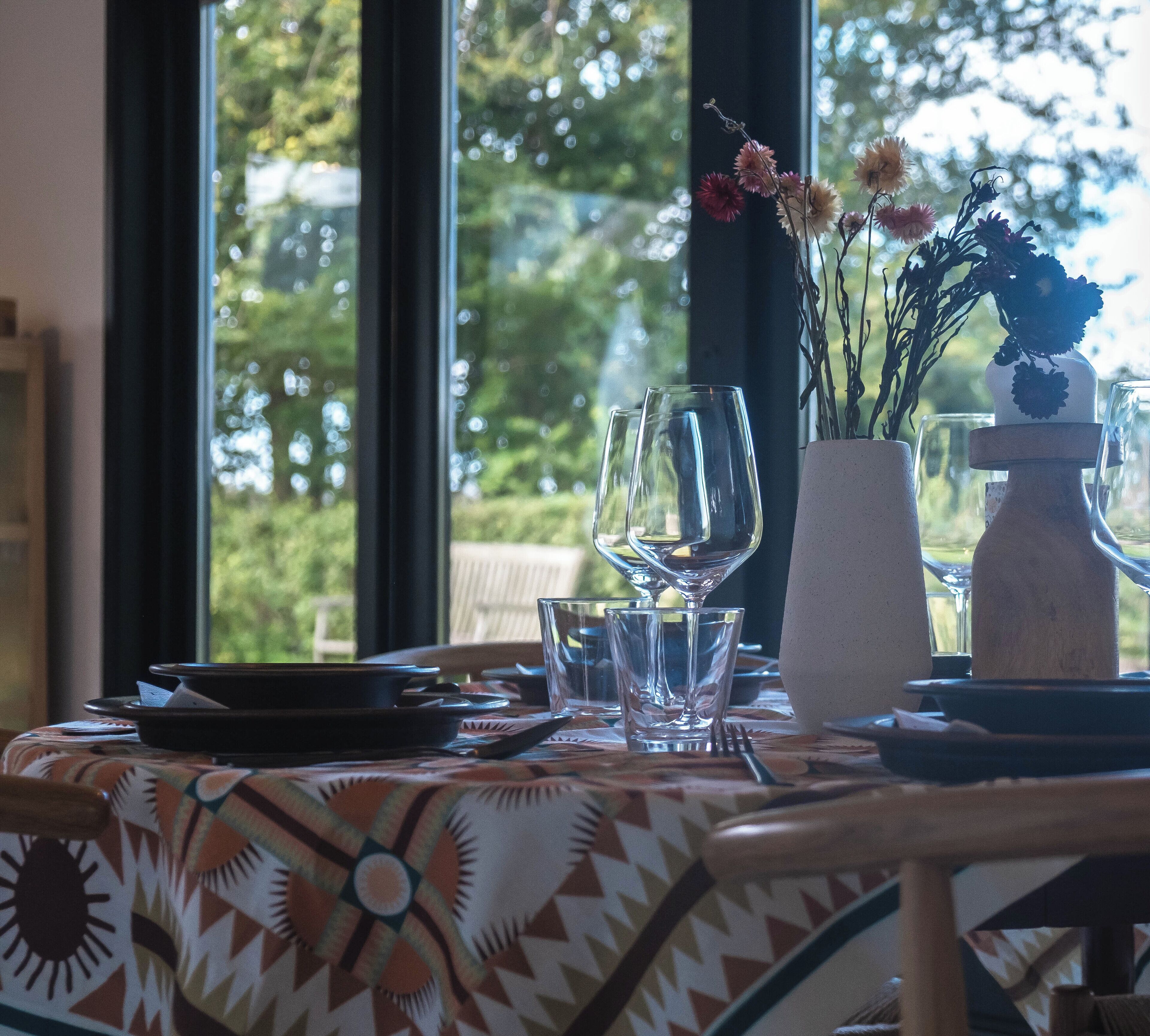 Une table à manger recouverte d’une nappe à motifs, deux verres à vin, un vase avec des fleurs et un vase décoratif.