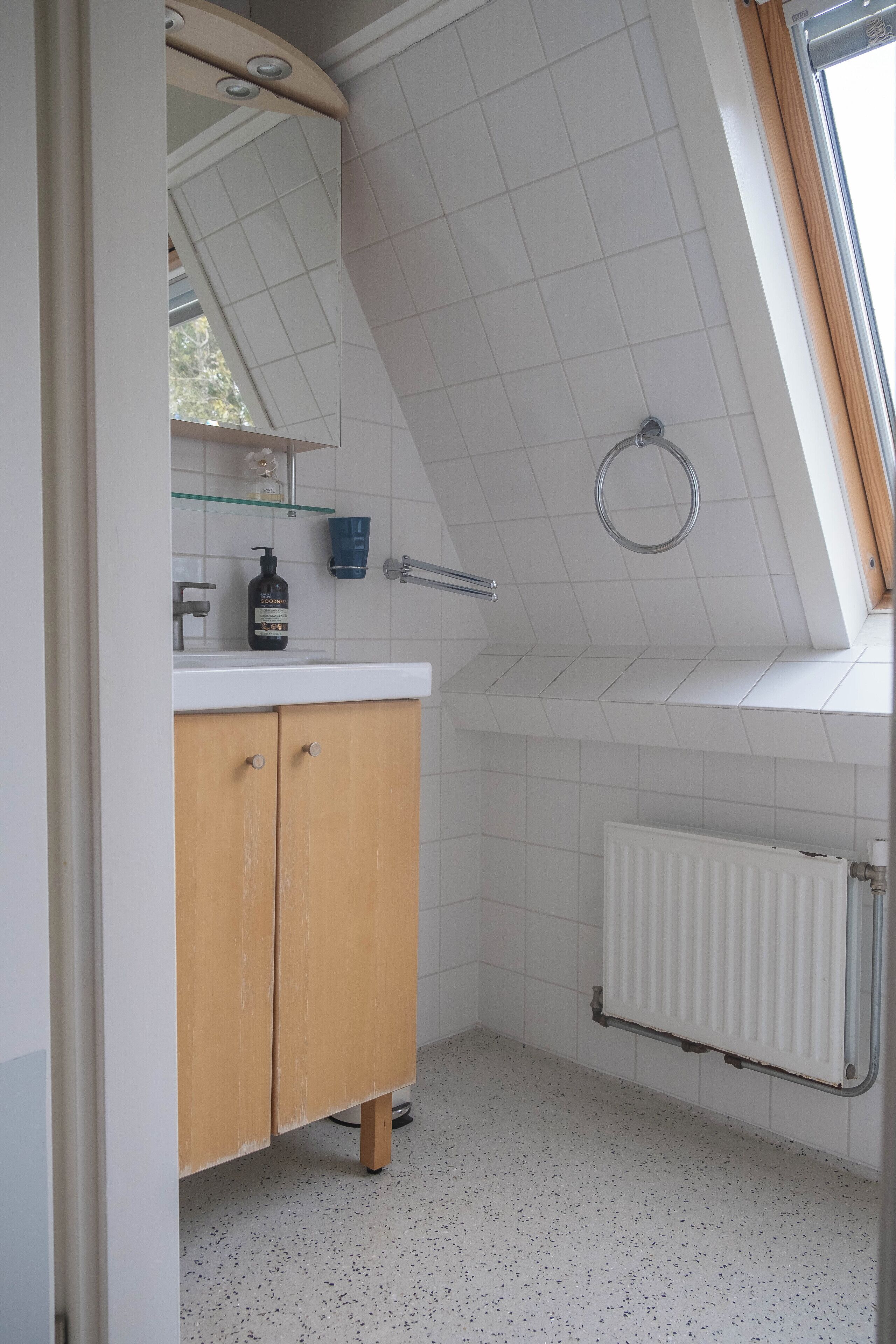 Une salle de bain avec un sol carrelé de carreaux blancs, un meuble en bois, un miroir, une étagère en verre, un radiateur blanc et une lucarne.
