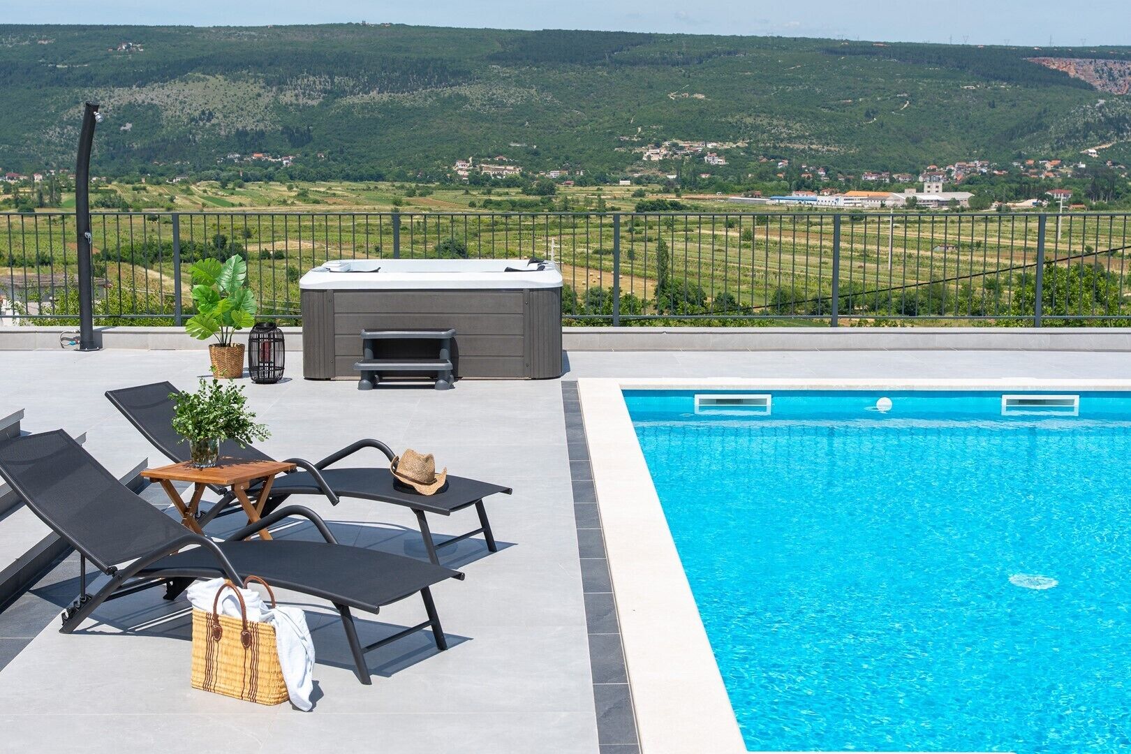 En swimmingpool med liggestole og et boblebad, omgivet af et hegn og udsigt til et bakket landskab.
