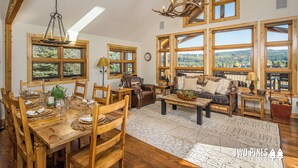 A spacious living area with a wooden dining table set for six, a leather sofa, and a large window offering a scenic view. - Enchanting Home! 10 Min Drive to Resort, 5 Min to Town Center! (Big Sky)