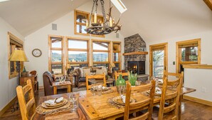A spacious dining area with a large wooden table set for six, a stone fireplace, and a view of the forest through large windows. - Enchanting Home! 10 Min Drive to Resort, 5 Min to Town Center! (Big Sky)