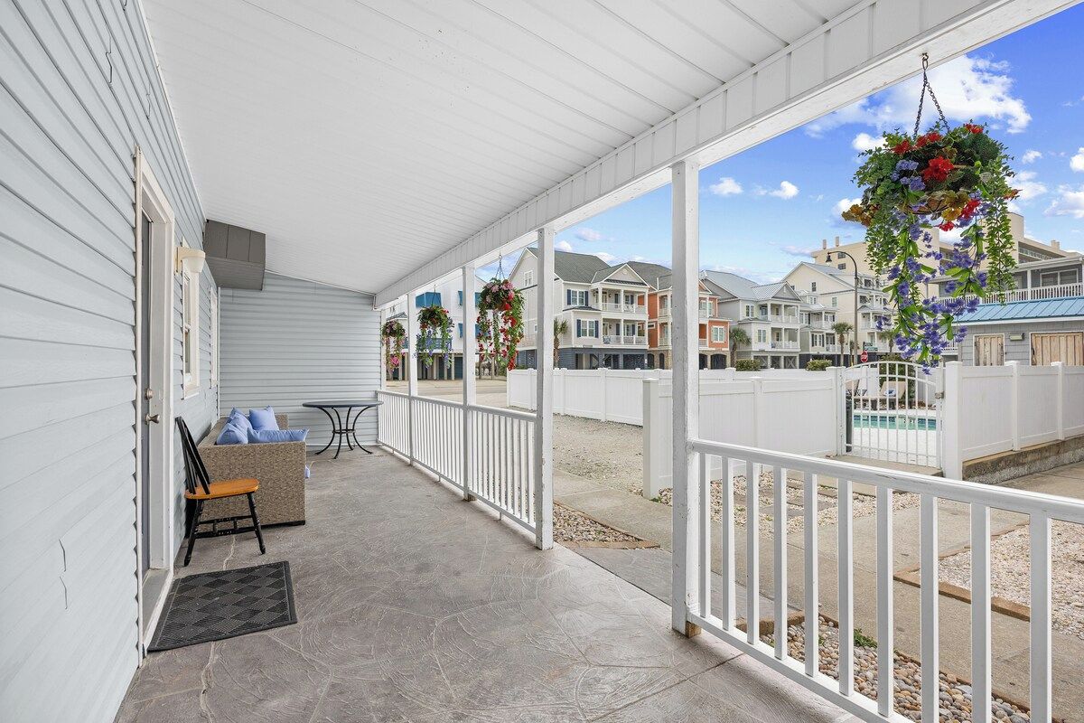 A covered porch with a seating area, a hanging flower basket, and a view of neighboring houses.